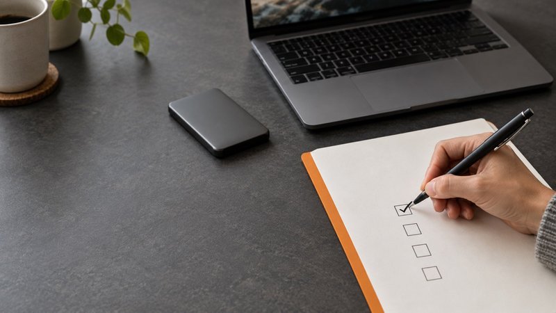 A person marks a paper checklist beside a laptop and an unconnected external drive.