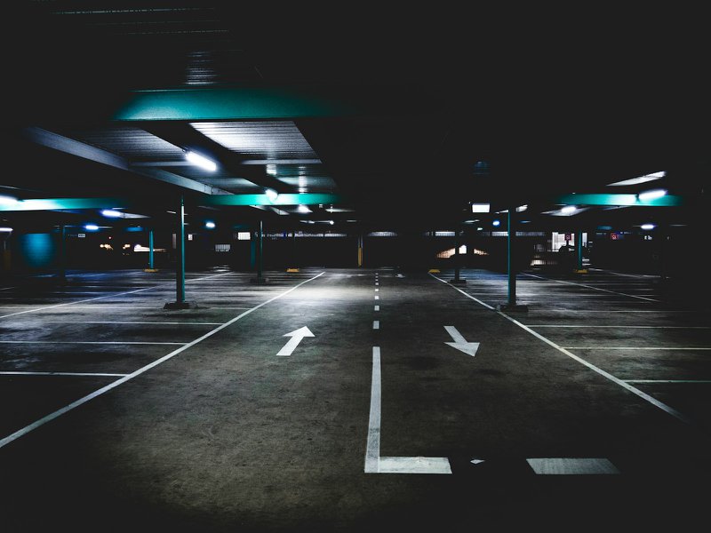 A dark, empty multi-storey car park with white directional arrows painted on the concrete floor and rows of vacant bays under strip lighting.