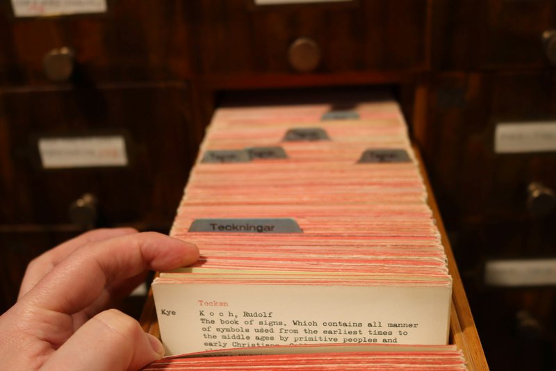 A hand pulling typed index cards from a wooden library card catalogue drawer, with rows of labelled brass-handled drawers behind.