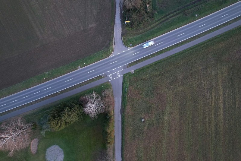 An aerial view of a crossroads where a main road meets a side lane between ploughed fields, a single car blurred with motion, muted gray-green tones.