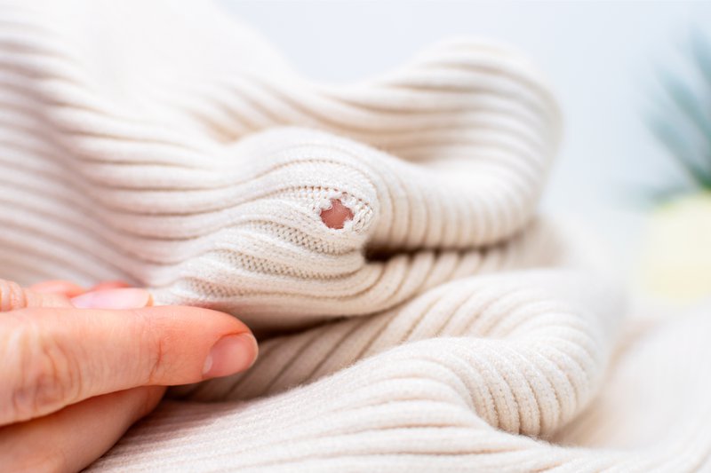 Clothing repair, craft sewing and textile recycle. Woman hands holding light knitted texture cloth with visible hole made by moth or ripped, close-up. Tailor holding damaged fabric with loose loops.