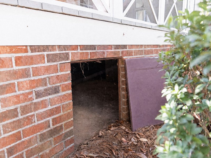 Open crawl space access door on a brick home, showing the dark unsealed space beneath the house.