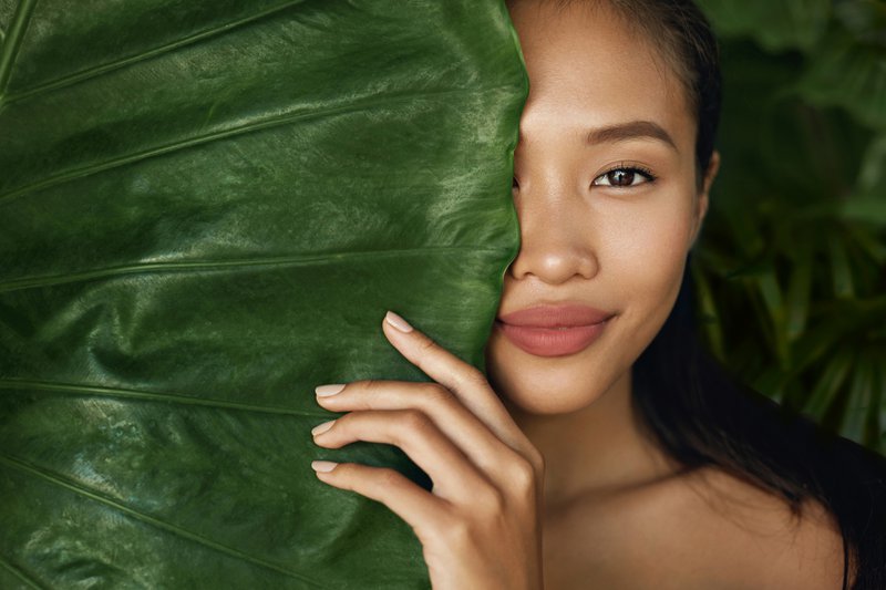 Rostro de belleza. Mujer modelo con maquillaje natural y piel sana detrás de planta de hojas verdes. Retrato de hermosa chica asiática con uñas desnudas, labios grandes y sonrisa sexy en la naturaleza tropical.