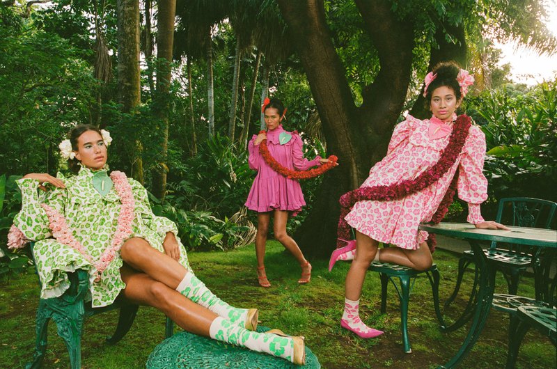 Three models in pink and green floral Rocket Ahuna designs with lei posed in a lush Hawaiian garden
