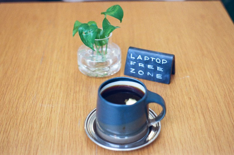 A cup of black coffee on a saucer beside a small plant in a glass jar and a black sign reading “Laptop Free Zone” on a wooden table.