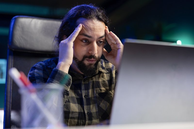Tired man rubbing his temples while looking at his laptop on desk. Frustrated male individual with his hands on forehead is working at home reviewing online documents on digital device.