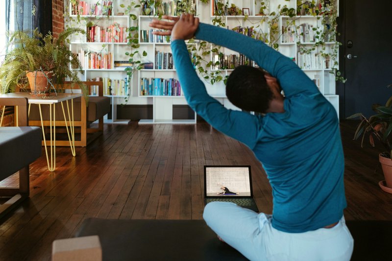 Man in a blue long-sleeved top sitting cross-legged on a yoga mat, stretching sideways with his arms overhead while following an online yoga class on a laptop