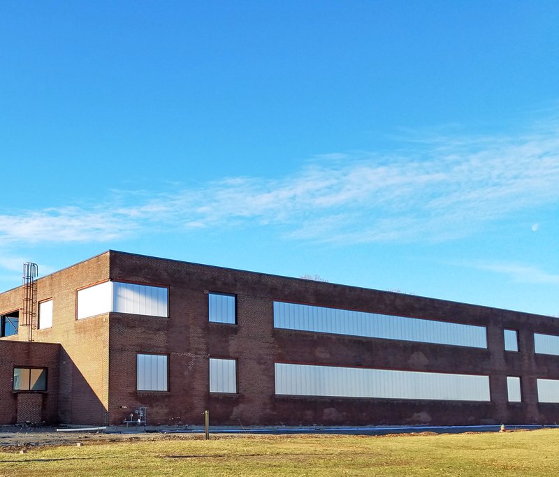 A long, two-story brick industrial building with several rectangular window openings filled with white, translucent panels. The building sits next to a grassy field under a wide, clear blue sky with thin, wispy clouds.