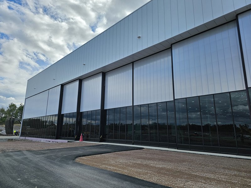 A large, modern industrial building featuring a facade of dark-tinted glass windows on the lower level and tall, translucent white panels above. The building sits next to a fresh asphalt path and a gravel construction site under a bright, cloudy sky.