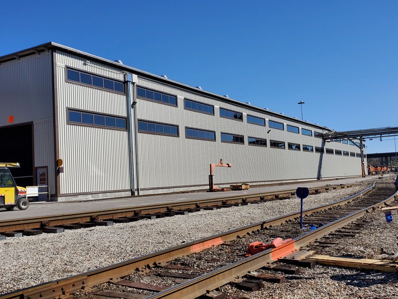 A long, corrugated metal industrial warehouse alongside a railyard, featuring two parallel rows of dark-framed ribbon windows stretching across the facade under a clear blue sky.