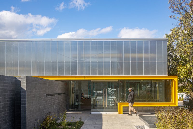 The exterior of the Animal Care Centers of NYC, featuring a vibrant yellow-framed glass entryway beneath a large, translucent ribbed facade. A man walks toward the entrance, which is flanked by a grey stone wall and set against a bright, partly cloudy sky.