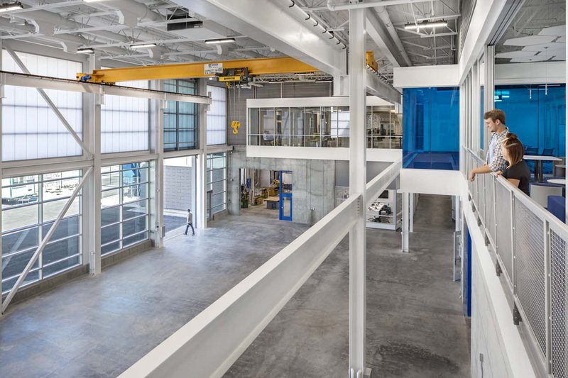 A high-angle view of a vast, industrial innovation lab or workshop. Two people stand on a second-floor balcony overlooking a wide concrete floor equipped with a large yellow overhead crane. The space features white steel beams and a massive wall of translucent light-diffusing panels that brighten the interior.