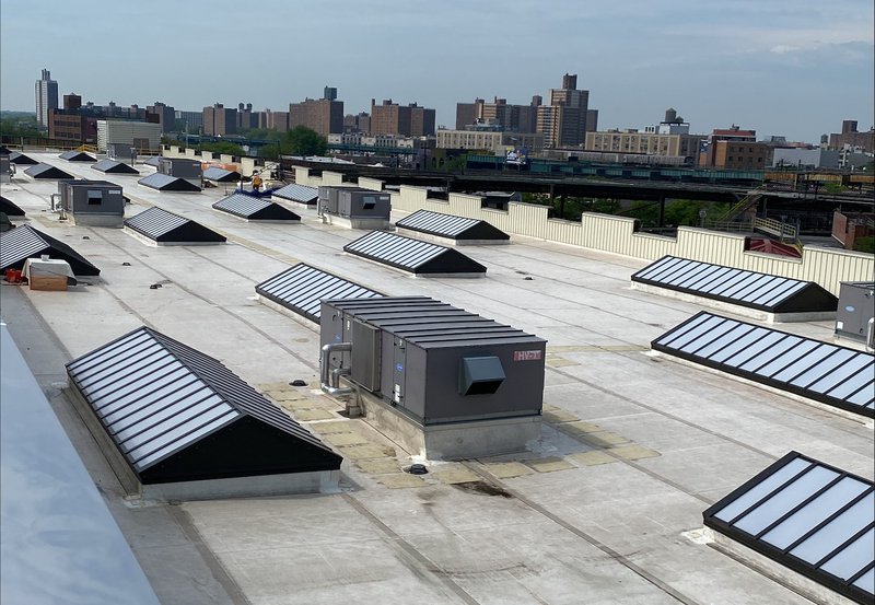 A wide-angle view of a large flat rooftop in an urban area, featuring rows of ridgetop skylights with black frames and translucent panels. Several industrial HVAC units are positioned among the skylights, with a cityscape of brick apartment buildings visible in the background under an overcast sky.