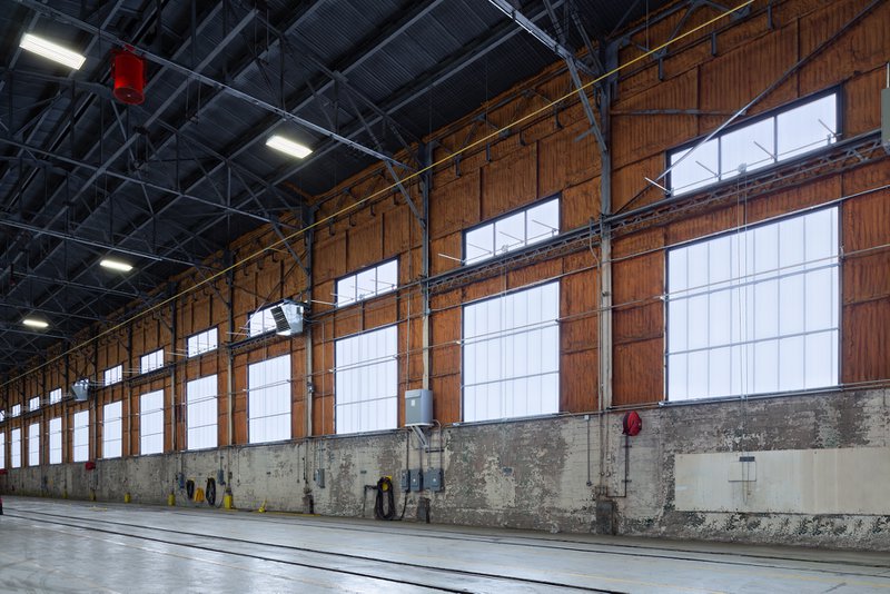 The interior of a Norfolk Southern Railyard facility, featuring a long wall of large, rectangular industrial windows with translucent white panels. The windows are set into a rustic wood-plank upper wall above a weathered concrete base, providing bright natural light to the expansive hangar space.