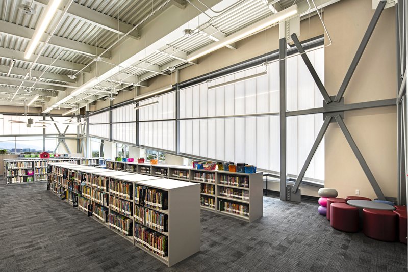 The modern interior of the Red Rock Elementary School library, featuring rows of low bookshelves and a circular red seating area. The space is illuminated by a massive wall of translucent white light-diffusing panels integrated into an industrial grey steel support structure.