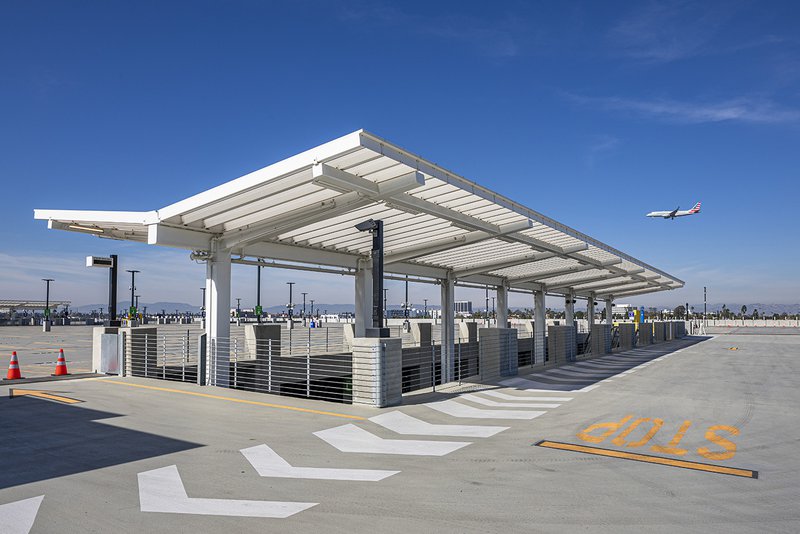 An airport parking garage rooftop featuring a long white metal canopy, with an American Airlines plane flying in the clear blue sky above the tarmac.