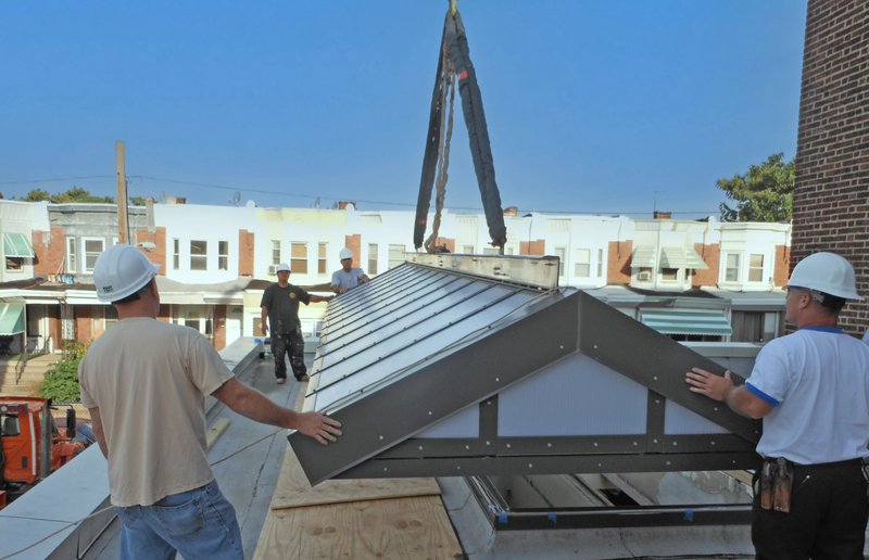 Construction workers using a crane to carefully install a large gabled skylight on a building’s rooftop against an urban backdrop.