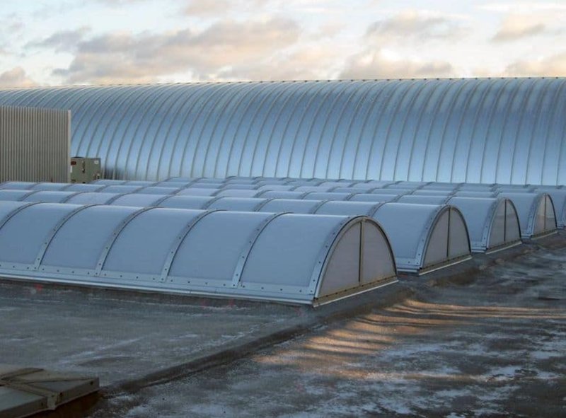Rows of barrel-vault industrial skylights with translucent panels and metal frames installed on a flat concrete rooftop, positioned in front of a large, curved corrugated metal building under a cloudy sky.