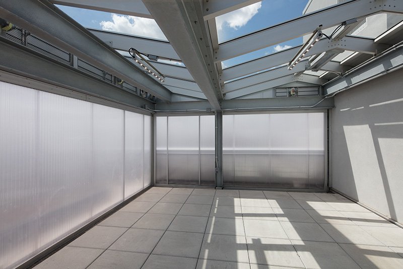 An empty outdoor terrace or balcony featuring translucent, ribbed wall panels and a modern grey steel support structure. The space is partially covered by a glass skylight roof that reveals a blue sky with white clouds, casting geometric shadows across the light-colored stone floor tiles.