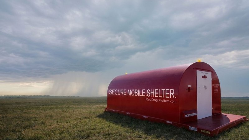 A Red Dog mobile storm shelter stands ready in an open field as a dark storm front and rain shaft approach in the background.