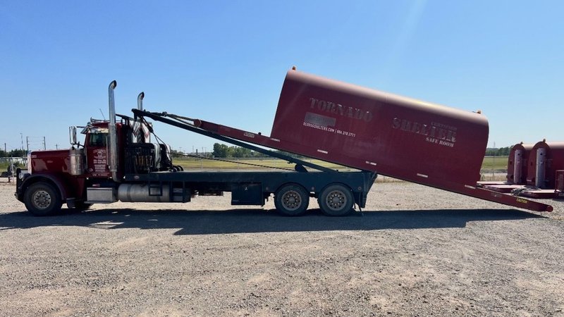 A truck is unloading a Red Dog storm shelter with the words "TORNADO SHELTER SAFE ROOM" written on its side.
