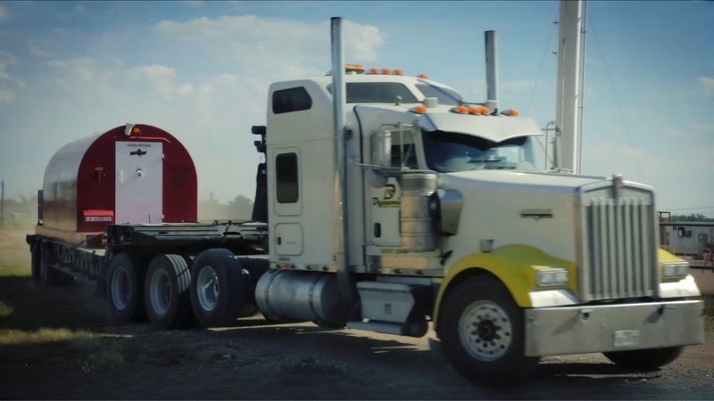 A white semi-truck with yellow accents on the hood transports a Red Dog mobile shelter unit on a flatbed trailer.