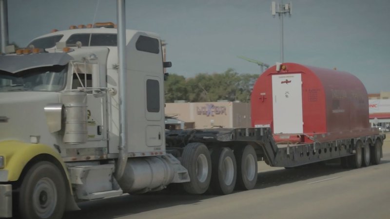A semi-truck transporting a red dog shelter on a flatbed trailer, offering a mobile alternative to in-ground storm shelters.