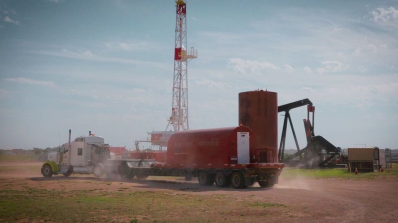 A red dog  storm shelter unit featuring specialized storm shelter ventilation is being transported to a facility with a drilling rig in the background.