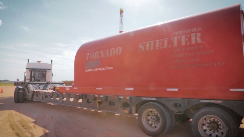 A semi-truck transporting a large red dog shelter on a flatbed trailer, delivering Modular Infrastructure Solutions.