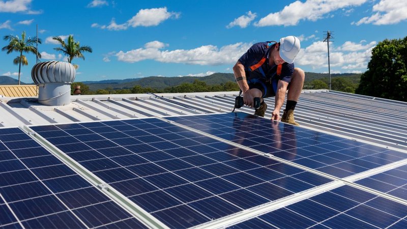 A worker installing panels on a metal roof, adhering to Solar Farm Construction Safety.