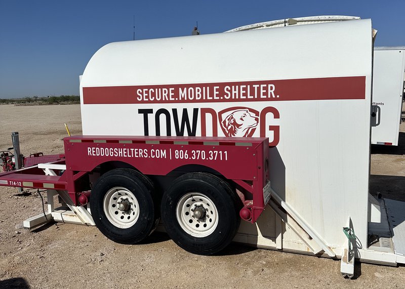 A side view of a white and red TOWDOG mobile storm shelter by Red Dog Shelters, parked on a dirt industrial site under a clear blue sky.
