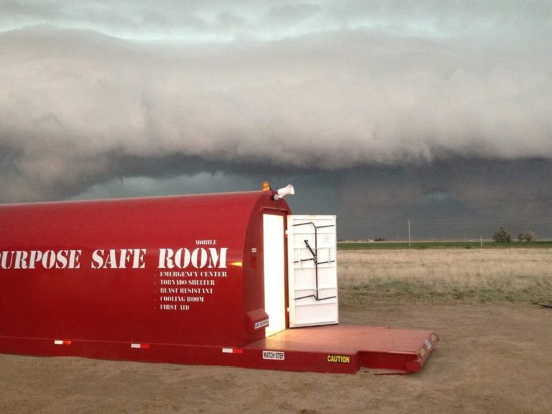The shelter sits on a flat, open field under an ominous, dark shelf cloud and a developing storm. Text on the side of the unit identifies it as an Emergency Center, Tornado Shelter, and Blast Resistant unit.