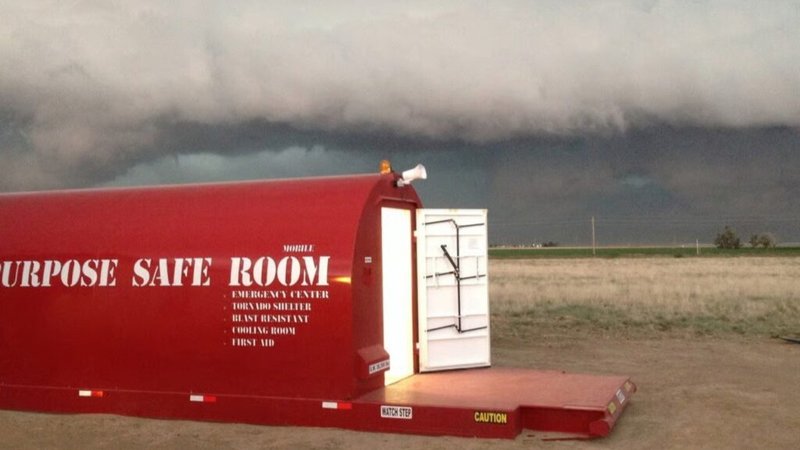 The shelter sits on a flat, open field under an ominous, dark shelf cloud and a developing storm. Text on the side of the unit identifies it as an Emergency Center, Tornado Shelter, and Blast Resistant unit.