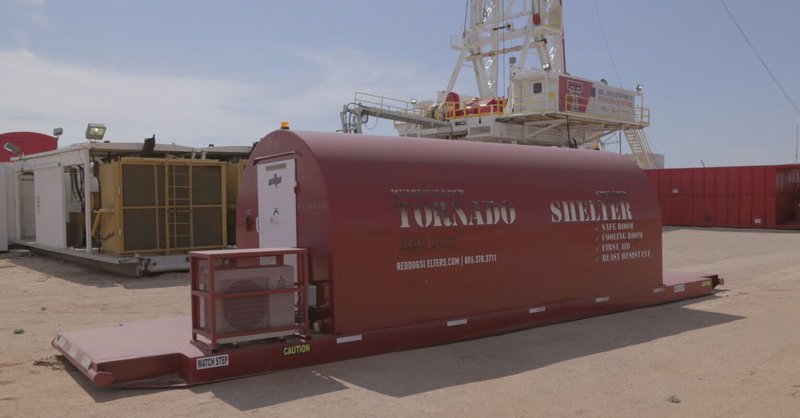 A red, industrial-grade Red Dog tornado shelter sits on a heavy-duty steel skid at a dusty outdoor job site with a drilling rig in the background.