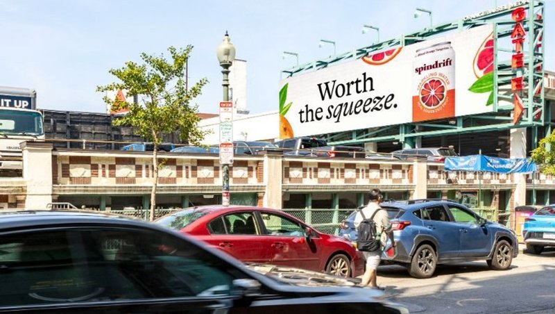 A large roadside billboard of a popular drink with text Worth the squeeze near city traffic.