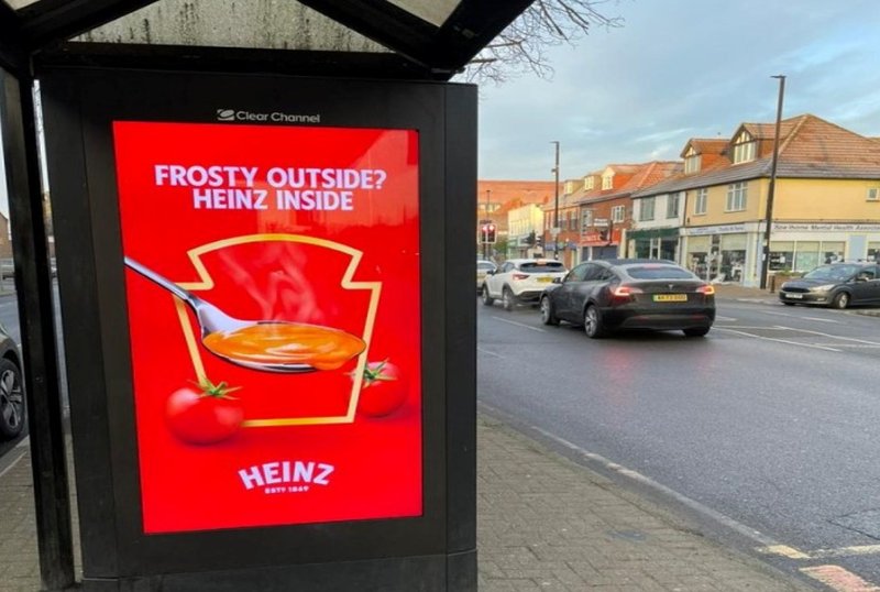 Street-level ad display of a popular ketchup brand on the side of a transit shed along a busy road