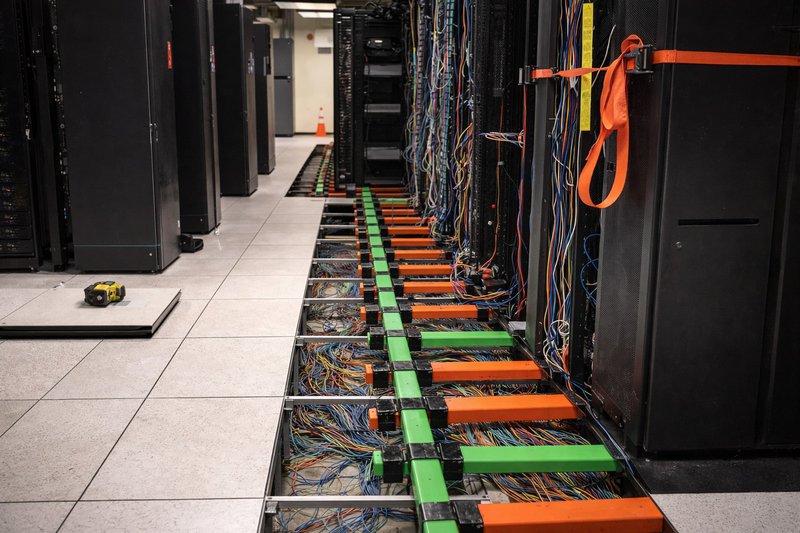 Wide view of a data center with raised floor panels removed, exposing organized cable trays and wiring beneath server racks during active infrastructure work.
