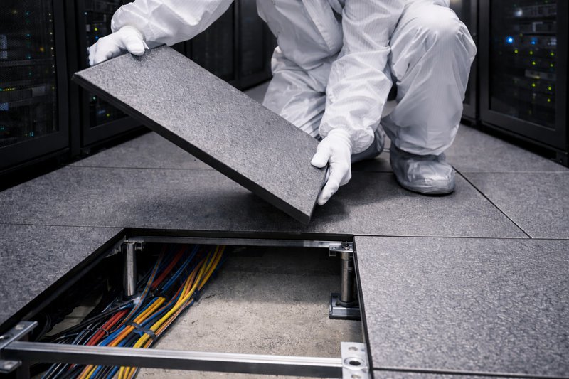 Technician lifting raised floor tile and using HEPA vacuum to clean subfloor plenum and cables in a live data center environment