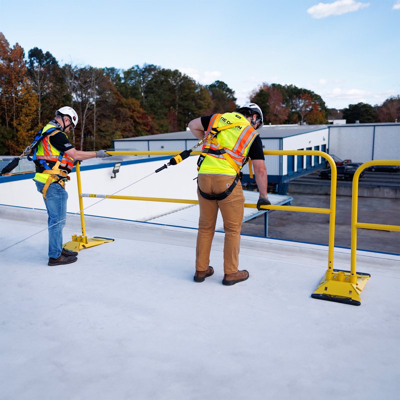 The image shows two workers, who are both wearing a headgear and a yellow vest, holding on to a guardrail. Both workers in the photo are wearing a safety harness for fall protection.