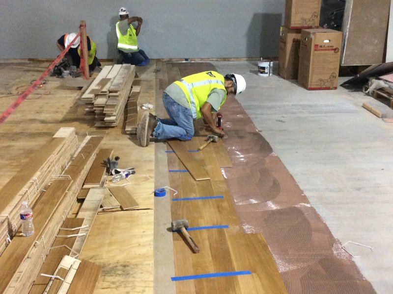 The image shows several people hard at work on a wood flooring installation project. They are using hammers to install the flooring.