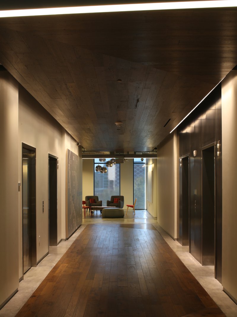 The image shows an interior that uses wood plank ceilings to hide the use of HVAC systems. Elevators, large windows, and couches can be seen in the image.