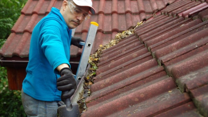 A homeowner clearing leaves from a roof gutter that lacks gutter guards.