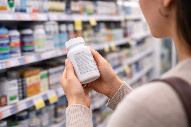 Horizontal image of a person holding a supplement bottle and examining the ingredient label in a store aisle with blurred shelves in the background.