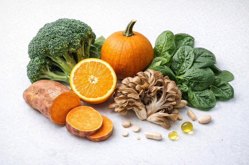 Assortment of whole food ingredients like broccoli, spinach, orange, and pumpkin used as natural vitamin sources.