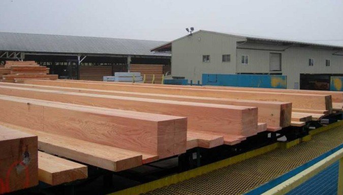 Large Douglas Fir timber beams arranged at an outdoor lumber facility near an industrial building.