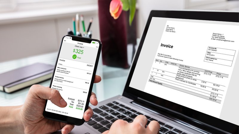 Person viewing a commercial invoice on a laptop while checking a paid invoice on a smartphone