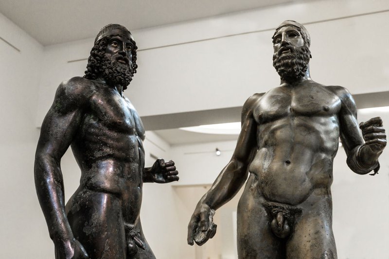 Deux statues antiques d'hommes nus en bronze dans un musée blanc.
