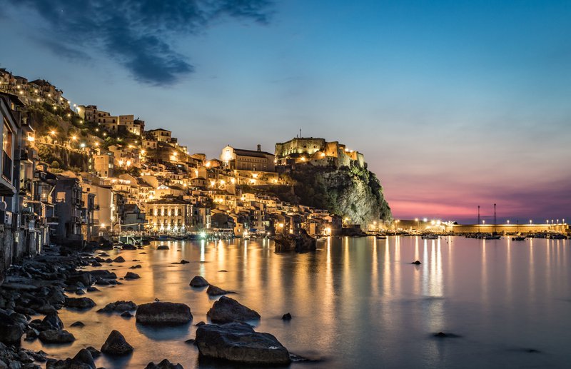 Photo de Scilla de nuit en Calabre, dans le sud de l'Italie. Au bord de l'eau, où les rochers dépassent de la surface. Et un beau ciel coloré et les lumières de la ville.