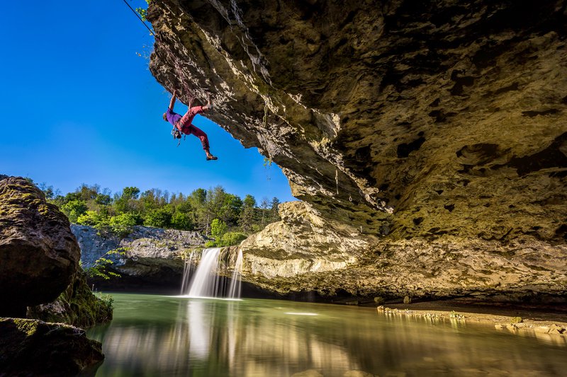 Klimmer op een hangende rots boven water met kleine waterval op de achtergrond