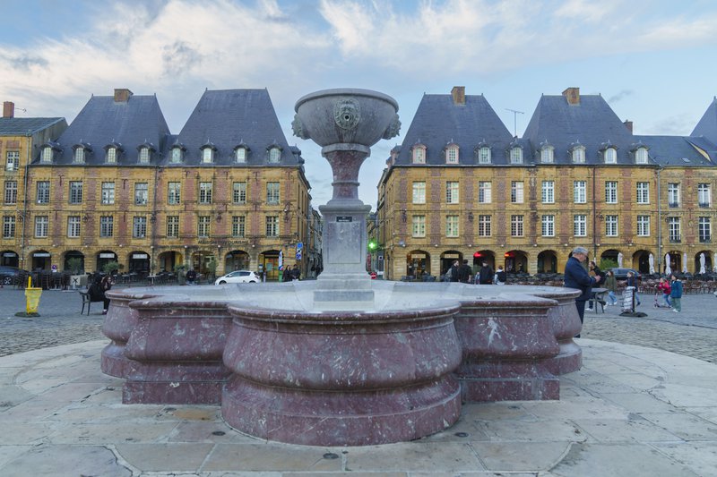 Fontaine imposante sur une place entourée de bâtiments ocres aux lignes épurées.